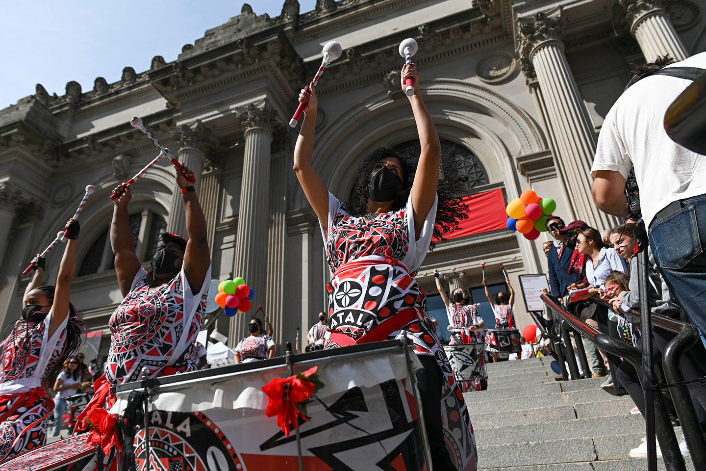 People in red, black and white outfits performing music to a crowd of people on the steps in front of The Metropolitan Museum of Art.